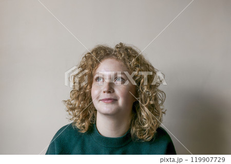 Young woman with curly, blonde hair gazes thoughtfully upwards, displaying a calm expression. Girl is dressed in a green sweater, surrounded by a neutral backdrop that enhances the moment. 119907729