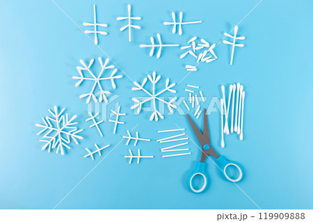 a crafting setup on a blue background. There are several cotton swabs arranged in the shape of snowflakes, with some swabs cut into smaller pieces. A pair of blue-handled scissors is also present, a crafting setup on a blue background. There are several cotton swabs arranged in the shape of snowflakes, with some swabs cut into smaller pieces. A pair of blue-handled scissors is also present, 119909888