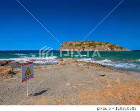 潮流注意の案内板が立つ砂浜と対岸にミノア文明遺跡の残る小島 (ギリシャ、クレタ島、モクロス) 119910603