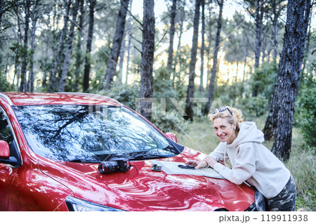 Woman orienting herself with a map and a compass close to a car in a forest. 119911938
