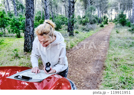 Woman orienting herself with a map and a compass close to a car in a forest. 119911951