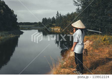 Samurai with katana in thought. A young samurai in traditional clothing and a conical hat stands by a river holding a katana, looking at a serene landscape, representing peace and tranquility. 119912224
