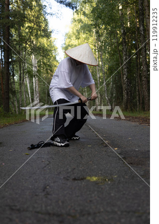 A focused samurai wearing an Asian hat practices with a katana in the forest embodying Japanese culture with skill and strength. The stance exudes the balance and strength of a warrior with a katana. A focused samurai wearing an Asian hat practices with a katana in the forest embodying Japanese culture with skill and strength. The stance exudes the balance and strength of a warrior with a katana. 119912235