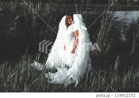 Cute angel standing in tall grass. Brunette with big white angel wings stands in a field of tall grass next to a lake on a sunny day, creating a serene and ethereal scene. Cute angel standing in tall grass. Brunette with big white angel wings stands in a field of tall grass next to a lake on a sunny day, creating a serene and ethereal scene. 119912252