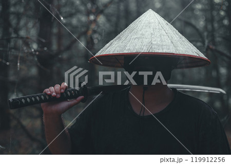 Majestic warrior with katana in the forest under pouring rain. Mysterious man in black mask and conical hat stands in the forest holding katana on shoulder on rainy day. 119912256