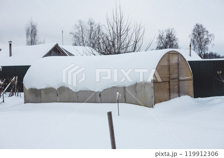 An old greenhouse stands in the backyard of a country house, covered with a thick layer of snow in a cold winter. Beautiful gentle winter in the garden with a covered greenhouse. An old greenhouse stands in the backyard of a country house, covered with a thick layer of snow in a cold winter. Beautiful gentle winter in the garden with a covered greenhouse. 119912306