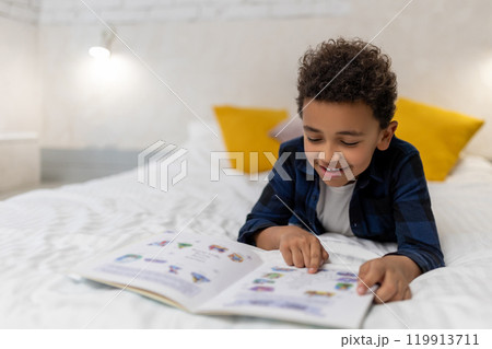 Curly-haired african american kid lying on the bed with a book and looking excited Curly-haired african american kid lying on the bed with a book and looking excited 119913711