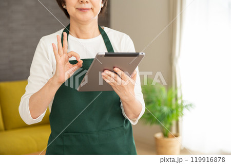 家のリビングでタブレットを見て丸サインをする緑のエプロンをしたシニア女性の主婦・家政婦 119916878