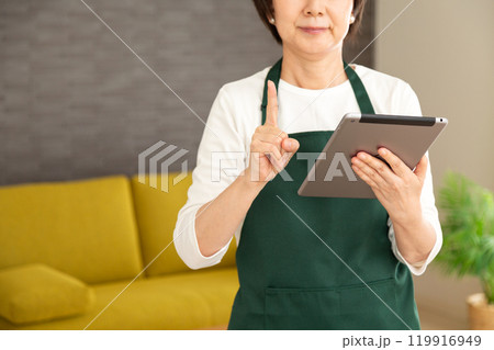家のリビングでタブレットを見て人差し指を立てる緑のエプロンをしたシニア女性の主婦・家政婦 家のリビングでタブレットを見て人差し指を立てる緑のエプロンをしたシニア女性の主婦・家政婦 119916949