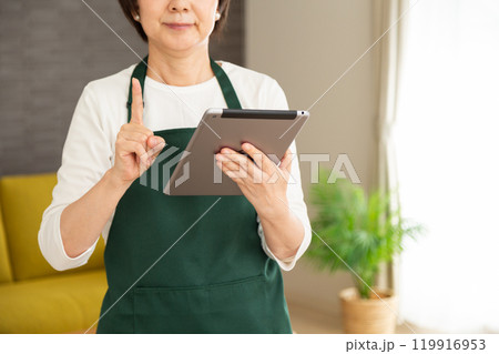 家のリビングでタブレットを見て人差し指を立てる緑のエプロンをしたシニア女性の主婦・家政婦 119916953
