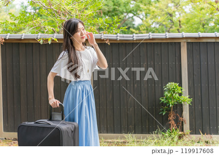 黒い塀の前の石畳の上をキャリーケースを引いて歩く若い女性旅行者 119917608