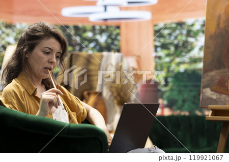 Female painter searching for new ideas and inspiration to complete a new painting masterpiece, using watercolor paint on canvas in her artistic workshop. Doing research online to get inspired. 119921067