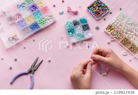 Creating colorful beaded jewelry with assorted beads and crafting tools on a light pink surface Creating colorful beaded jewelry with assorted beads and crafting tools on a light pink surface 119922936