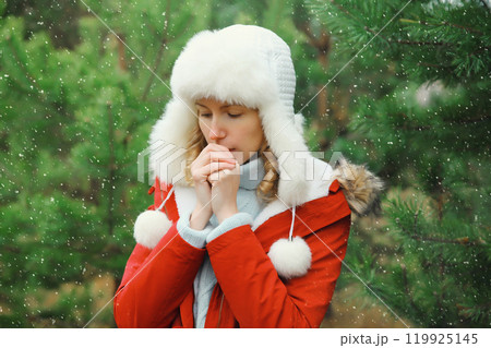 Woman is cold, freezing trying to warm up outdoors in winter park against Christmas tree in forest Woman is cold, freezing trying to warm up outdoors in winter park against Christmas tree in forest 119925145