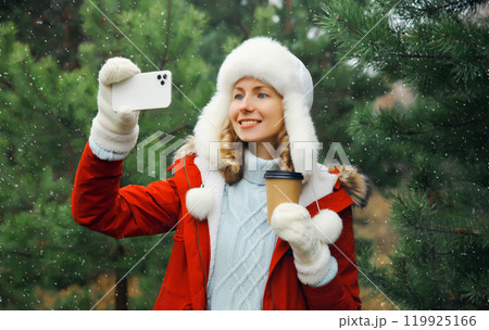 Happy smiling woman taking selfie with smartphone in winter park against Christmas tree, using phone 119925166