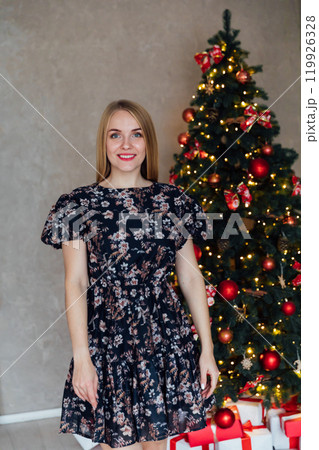 Blonde woman in dress smiling with gifts at Christmas tree for new year 119926328