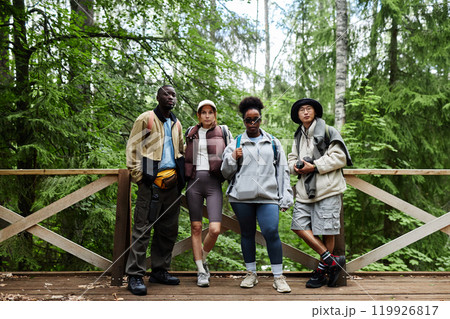 Full length portrait of multiethnic group of young people as tourists in nature looking at camera standing on bridge with beautiful scenery in background Full length portrait of multiethnic group of young people as tourists in nature looking at camera standing on bridge with beautiful scenery in background 119926817