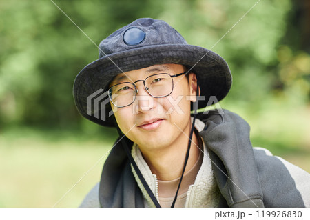 Close up portrait of kind young Asian man wearing hat and looking at camera while hiking in nature against blurred greenery background, copy space 119926830