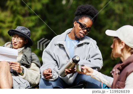 Multiethnic group of young tourists taking break outdoors in nature with smiling Black woman poring hot tea to friend Multiethnic group of young tourists taking break outdoors in nature with smiling Black woman poring hot tea to friend 119926839
