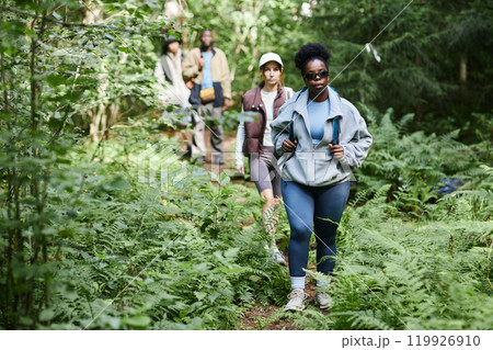 Multiethnic group of tourists walking in row on forest path while hiking together, focus on Black woman in front Multiethnic group of tourists walking in row on forest path while hiking together, focus on Black woman in front 119926910