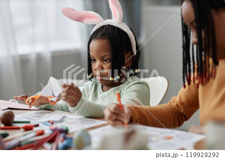 Focused child wearing bunny ears headband and holding scissors while making craft at table adult assisting with arts and crafts 119928292