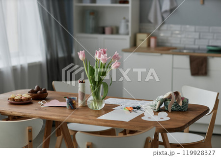 Wooden dining table adorned with Easter crafts and decorated eggs, bright tulips in glass vase adding freshness to the room, kitchen background with white cabinets visible Wooden dining table adorned with Easter crafts and decorated eggs, bright tulips in glass vase adding freshness to the room, kitchen background with white cabinets visible 119928327