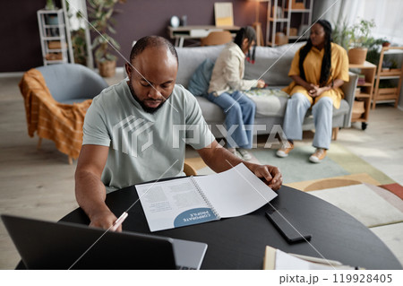 Man sitting at table working on laptop while family members engage in activities in living room with modern decor, creating a lively atmosphere Man sitting at table working on laptop while family members engage in activities in living room with modern decor, creating a lively atmosphere 119928405