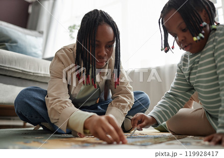 Two African American children sitting on floor assembling puzzle in cozy living room setting, showing concentration and teamwork, with natural light from window illuminating the scene 119928417