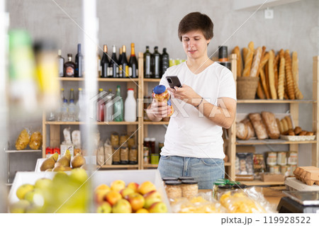 Young man in store chooses canned beans, scan product label with phone. Young man in store chooses canned beans, scan product label with phone. 119928522