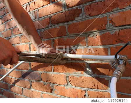 hands with long gas wrench tightening fastener on white plastic pipe coming out of brick wall, setting up outdoor water supply for garden irrigation or outdoor water use hands with long gas wrench tightening fastener on white plastic pipe coming out of brick wall, setting up outdoor water supply for garden irrigation or outdoor water use 119929144