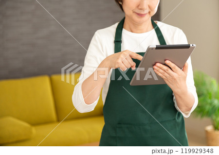 家のリビングでタブレットを見て微笑む緑のエプロンをしたシニア女性の主婦・家政婦 119929348