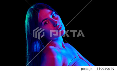 Portrait of young woman with long hair tilted head back and demonstrate her silky hair in neon light against black background. Portrait of young woman with long hair tilted head back and demonstrate her silky hair in neon light against black background. 119939015