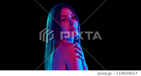 Portrait of young attractive woman posing in bright neon light, gently touching her shoulder against black studio background. 119939017