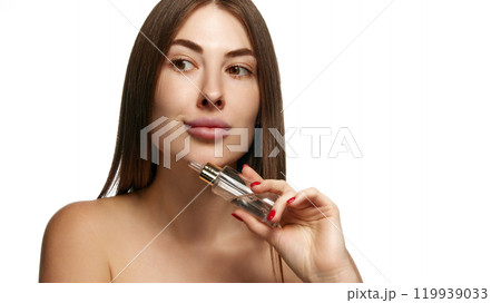 Pretty woman with bare shoulders holding serum bottle to her face for skincare against white background. Facial care treatment. 119939033