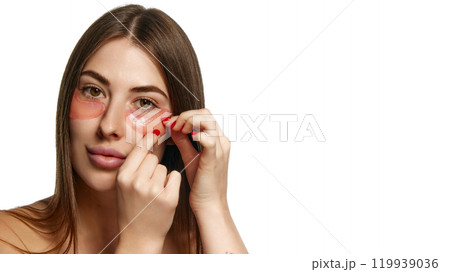 Woman with bare shoulders applying under-eye hydrogel patches for hydration against white background. With copy space. Woman with bare shoulders applying under-eye hydrogel patches for hydration against white background. With copy space. 119939036