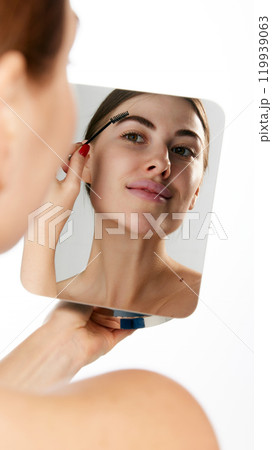Woman brushing her eyebrows while looking in mirror, highlighting precise grooming against white background. Woman brushing her eyebrows while looking in mirror, highlighting precise grooming against white background. 119939063