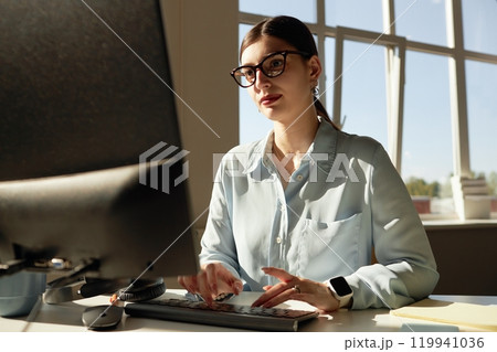Portrait of successful young businesswoman using computer enjoying workday in office lit by sunlight, copy space 119941036