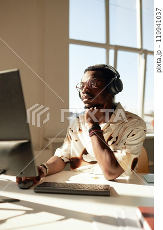 Vertical warm tone portrait of African American man enjoying workday in office lit by sunlight and using computer Vertical warm tone portrait of African American man enjoying workday in office lit by sunlight and using computer 119941037