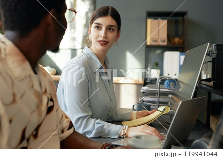 Two people working together in office, focus on smiling young woman talking to colleague while using computer 119941044