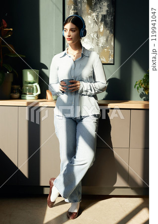 Vertical full length portrait of confident and elegant young woman wearing headphones and standing by coffee station in office lit by graphic sunlight 119941047
