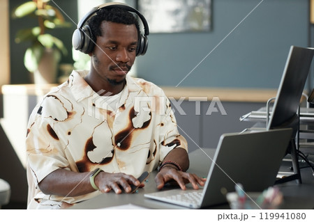 Portrait of trendy African American man using laptop and tech gadgets in office and wearing big headphones at modern workplace, copy space Portrait of trendy African American man using laptop and tech gadgets in office and wearing big headphones at modern workplace, copy space 119941080