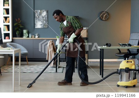 Full length side view shot of young African American man working in cleaning service and vacuuming floors in home or office interior, copy space Full length side view shot of young African American man working in cleaning service and vacuuming floors in home or office interior, copy space 119941166