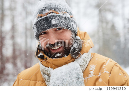 Portrait of bearded adult man smiling at camera covered in snow outdoors in winter forest with genuine emotion 119941210