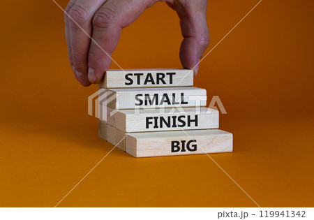 Start small finish big symbol. Concept words 'Start small finish big' on wooden blocks on a beautiful orange background. Businessman hand. Business, motivational and start small finish big concept. Start small finish big symbol. Concept words 'Start small finish big' on wooden blocks on a beautiful orange background. Businessman hand. Business, motivational and start small finish big concept. 119941342