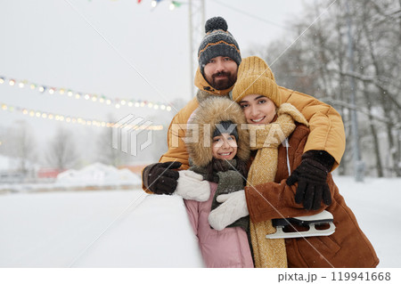 Waist up portrait of happy young family with daughter standing by skating rink in winter park together and looking at camera smiling copy space 119941668