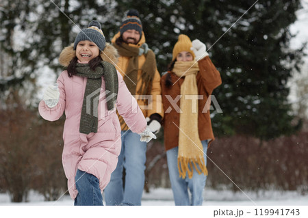 Candid shot of cute young girl running in snowy forest with parents in background happy family enjoying holidays outdoors copy space Candid shot of cute young girl running in snowy forest with parents in background happy family enjoying holidays outdoors copy space 119941743