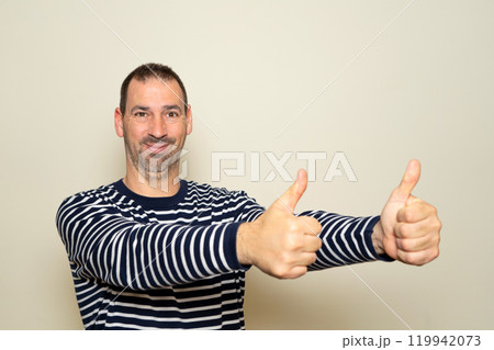 Bearded hispanic man in his 40s wearing a striped sweater with his arms outstretched and thumbs up in a sign of approval and positivity, isolated on a beige background. 119942073