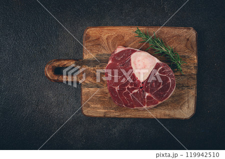 a shank on the bone, raw meat for ossobuco, on a wooden chopping board, with a branch of rosemary, top view, no people. a shank on the bone, raw meat for ossobuco, on a wooden chopping board, with a branch of rosemary, top view, no people. 119942510