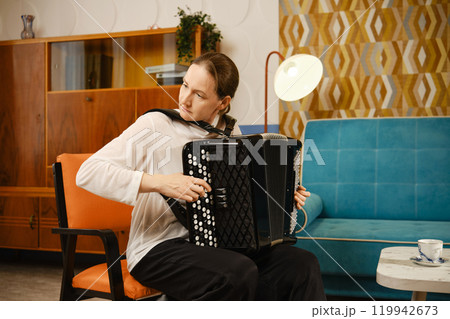 A middle-aged woman playing the accordion at home in a cozy living room setting during the afternoon A middle-aged woman playing the accordion at home in a cozy living room setting during the afternoon 119942673