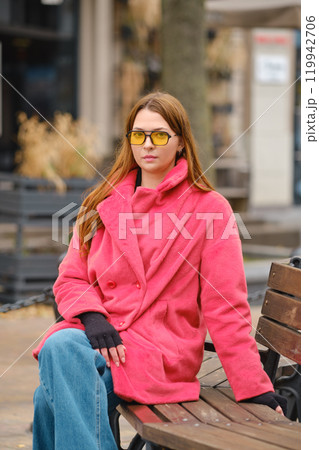 Young woman in pink coat and yellow sunglasses sitting on a bench in a city square during autumn 119942706